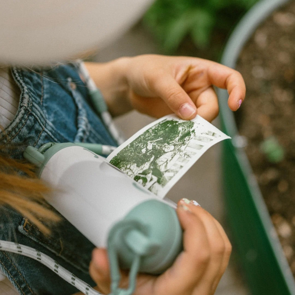 A close-up lifestyle shot of a person holding a mint green and white reusable water bottle while holding a printed photo strip. The person is wearing a denim jacket, and the background shows a blurred garden bed.