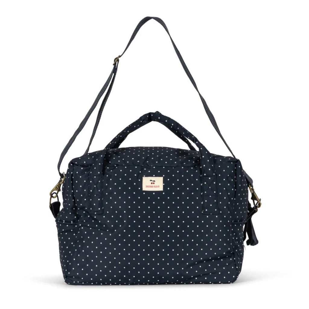 A navy blue fabric bag with a white polka dot pattern, featuring top handles, a detachable shoulder strap, and a small beige brand label on the front.