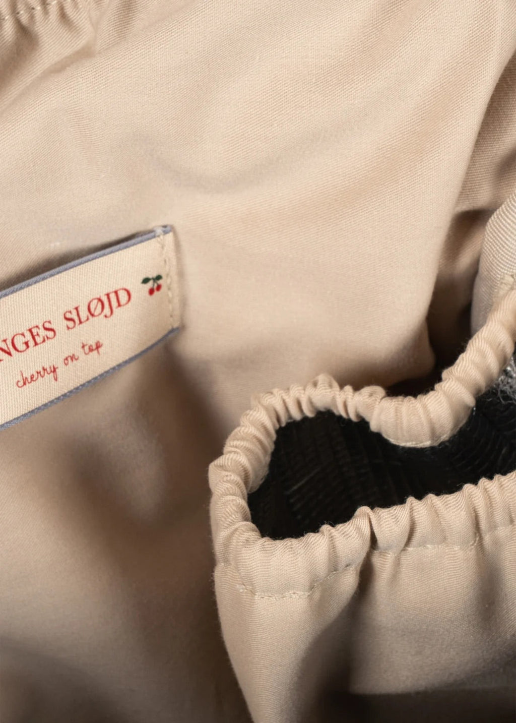 Close-up of a beige garment with gathered elastic trim and a sewn-in fabric label reading "NGES SLOJD" and "cherry on tap."