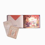 An open illustrated book or album with a warm-toned fantasy scene on the right page and printed cards or inserts placed in front, photographed on a clean white background.