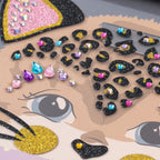 Close-up of a decorative sticker or decal sheet featuring a cartoon cat face with glittery black and gold details, pink and gold glitter patches, and scattered rhinestone embellishments in pink, blue, purple, and gold.