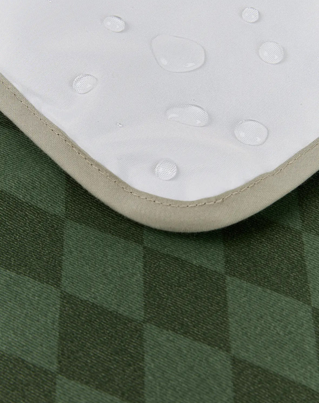 Close-up of a white waterproof mattress protector with water droplets on its surface, showing its beige stitched edge over a green plaid fabric background.