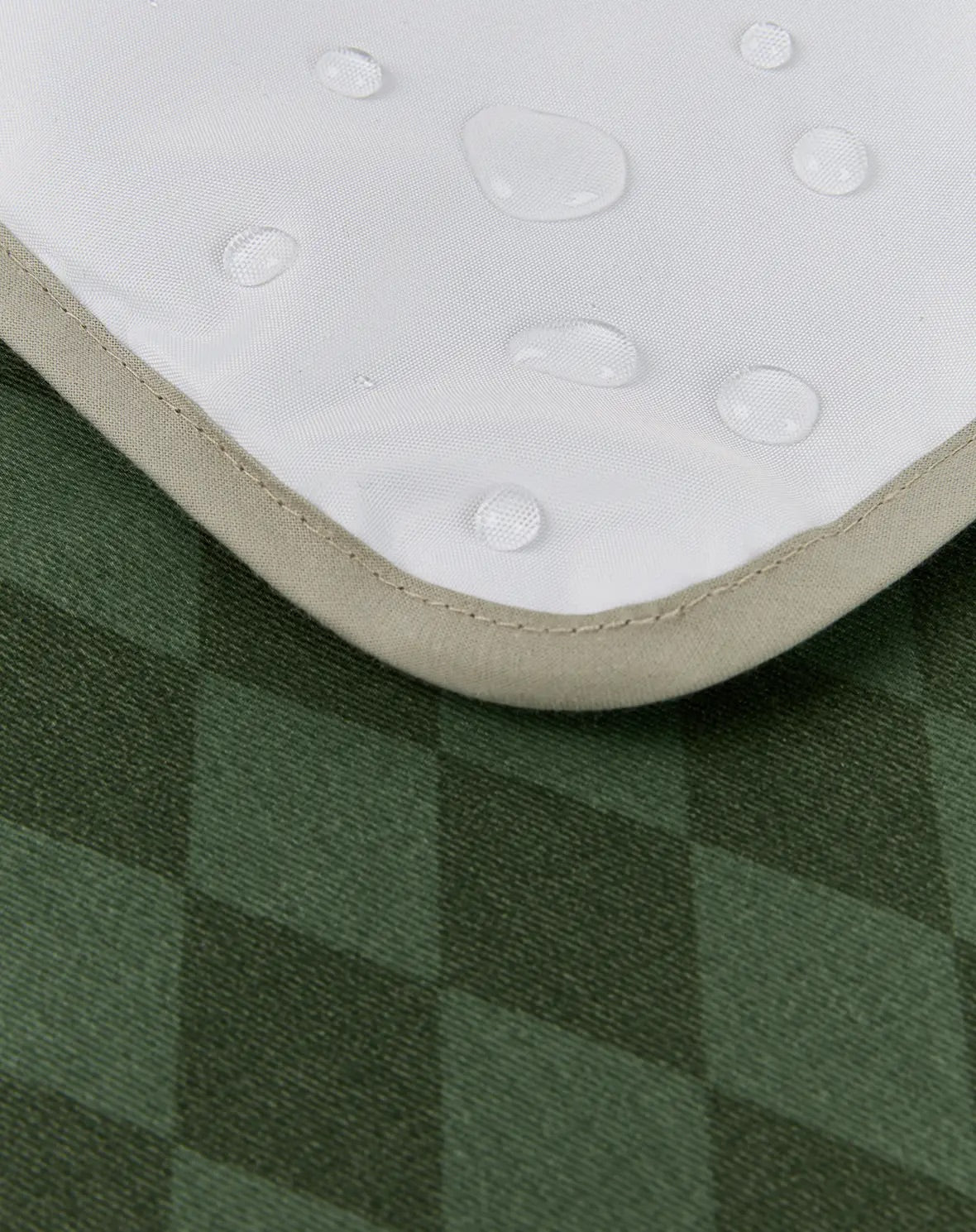 Close-up of a white waterproof mattress protector with water droplets on its surface, showing its beige stitched edge over a green plaid fabric background.