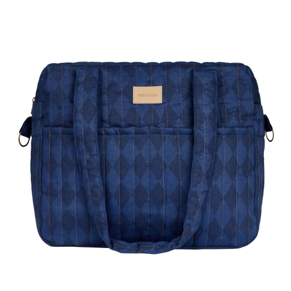 A navy blue quilted diaper bag with a subtle diamond pattern, featuring a tan leather brand patch on the front, set against a plain white background.