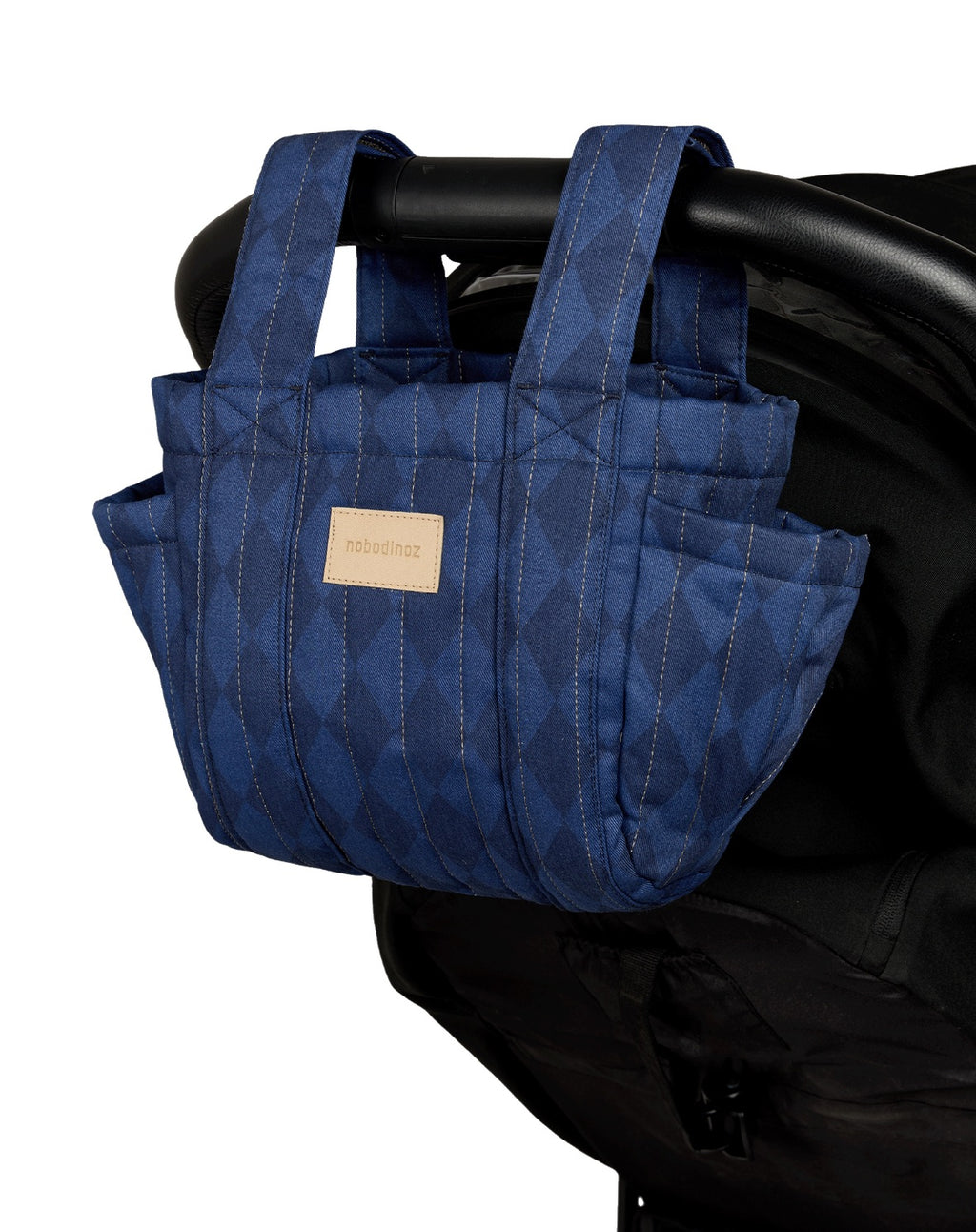 A blue quilted stroller bag with a subtle geometric pattern, hanging from a black stroller handle. The bag features multiple pockets and a small beige brand label on the front.