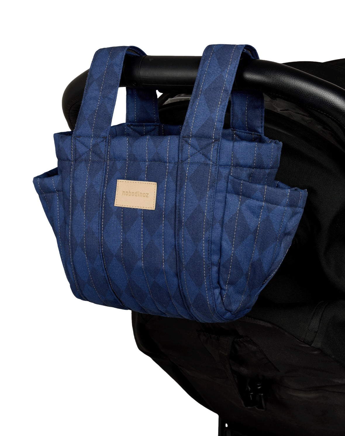 A blue quilted stroller bag with a subtle geometric pattern, hanging from a black stroller handle. The bag features multiple pockets and a small beige brand label on the front.